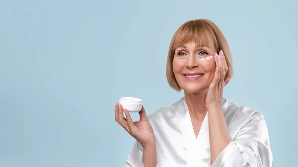 Smiling mature woman applying rejuvenating cream on her face over blue studio background, banner design with empty space — Stock Photo, Image