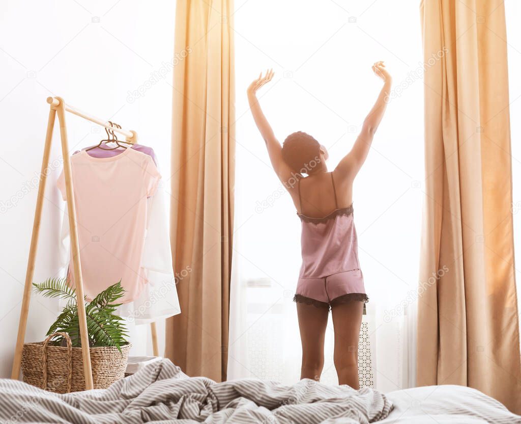 Smiling young african american lady awake after healthy sleep sitting in cozy modern comfortable bedroom interior