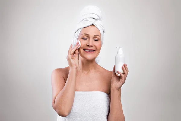 Senior woman wearing bath towels, enjoying her beauty and face care routine, applying skin tonic on light background — Stock Photo, Image