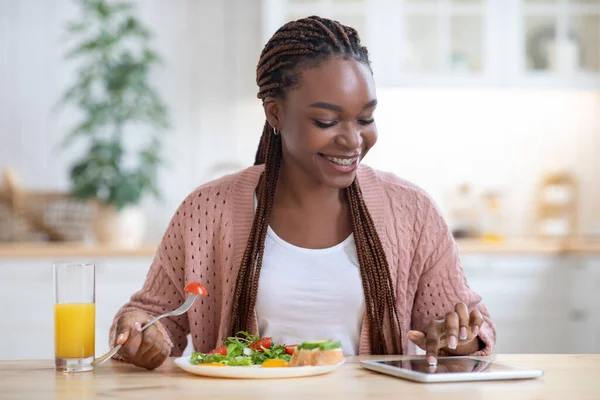 Sonriente joven mujer negra usando tableta digital mientras desayuna en la cocina —  Fotos de Stock