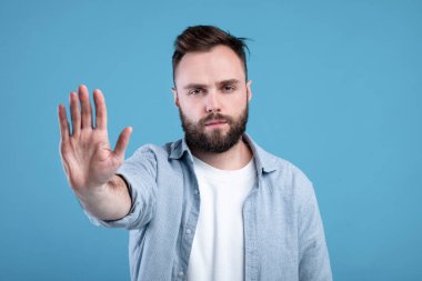 Uzak dur, mesafeni koru. Mavi stüdyo arka planında avuçtan kameraya DUR hareketi gösteren hoşnutsuz genç adam