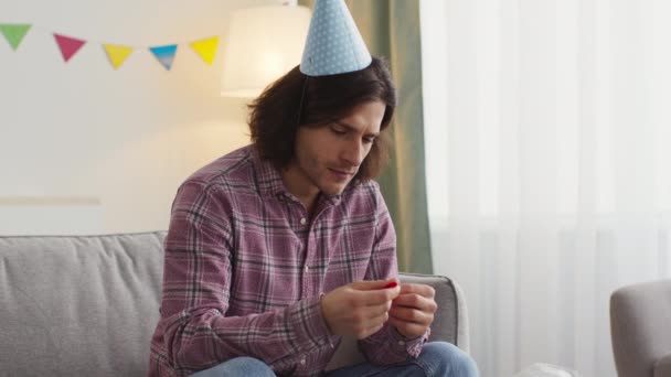 Zelfisolatie en eenzaamheid. Jonge ongelukkige verjaardagsman in feestmuts gooit ballon weg en kijkt droevig naar raam — Stockvideo