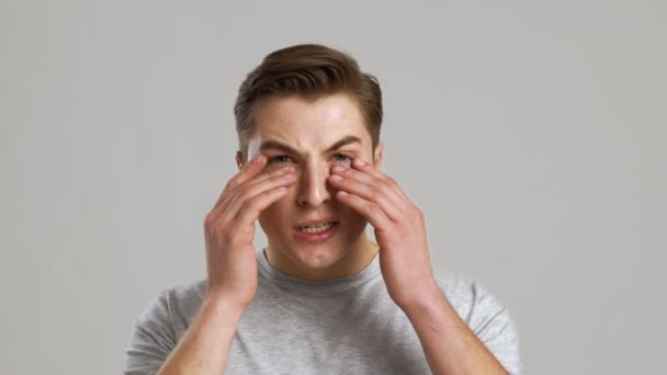 Problema de vista. Primer plano del joven frotando sus ojos inflamados, fondo gris del estudio — Vídeo de stock