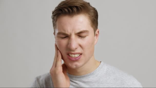 Diş ağrısı. Akut diş ağrısından muzdarip genç bir adam, ağrıyan yanağını ovuyor, gri stüdyo geçmişi var. — Stok video