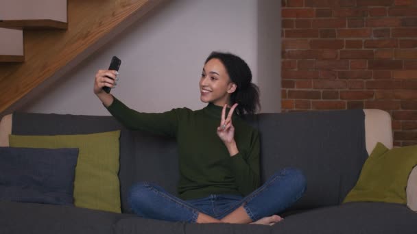 可爱的黑人女士在智能手机相机上自拍，在家里玩的开心，坐在沙发上，慢动作 — 图库视频影像