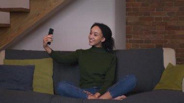Neşeli Afro-Amerikan kadın videosu arkadaşlarını arıyor, akıllı telefondan sohbet ediyor, evdeki kanepede oturuyor, ağır çekimde.