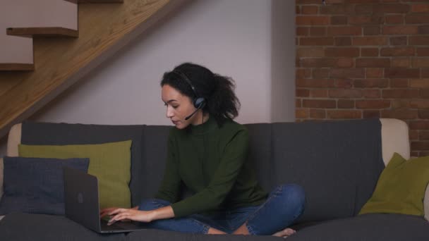 Hjälplinje hemma. Ung svart kvinna i headset prata med kunden och skriva på bärbar dator, arbeta på distans — Stockvideo