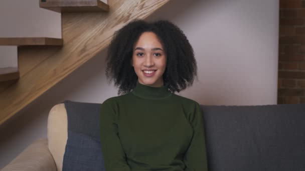 Retrato de cerca de la joven mujer negra sentada en el sofá en casa, sonriendo a la cámara — Vídeos de Stock
