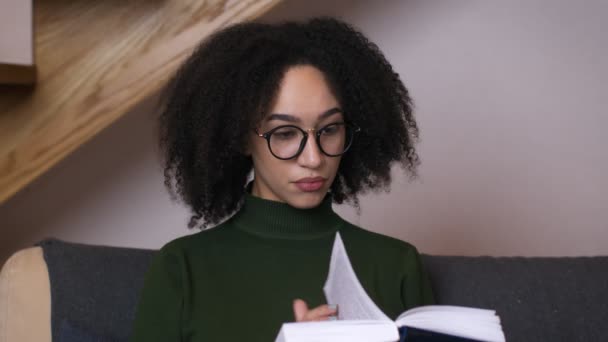 Joven mujer afroamericana en anteojos leyendo libro en casa interior, de cerca, cámara lenta — Vídeo de stock