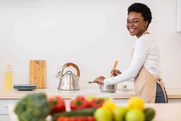 Счастливая африканская женщина готовит ужин на кухне, улыбаясь в камеру — стоковое фото