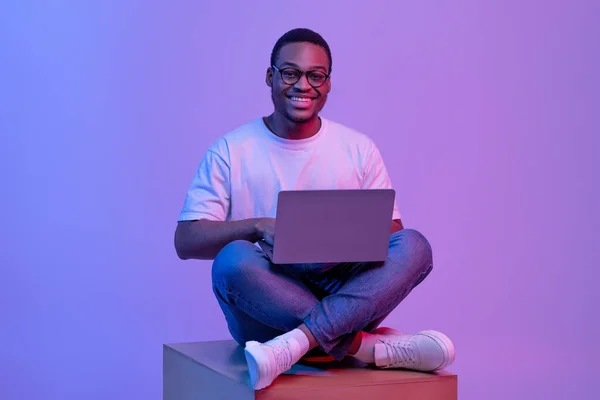 Happy Black Student Guy In Eyeglasses Sitting With Laptop In Neon Lighting