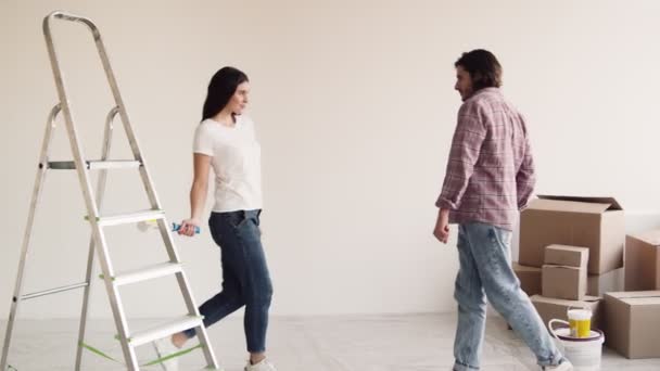 Vreugdevolle woningrenovatie. jong gelukkig paar het maken van reparatie, dansen samen met verfroller in leeg appartement — Stockvideo