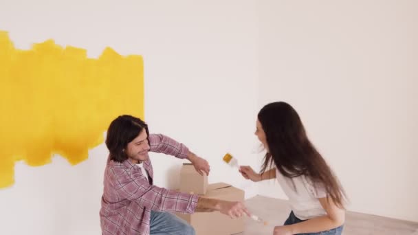 Curiosa reparación. Joven casado hombre y mujer pintando paredes en casa, tratando de pintarse, riendo y divirtiéndose — Vídeos de Stock