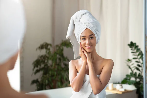 Mujer afroamericana milenaria satisfecha en toalla presiona las manos a la cara y mira el reflejo en el espejo en el baño — Foto de Stock