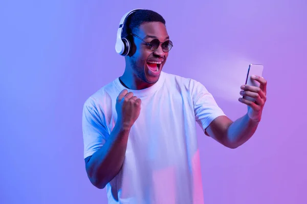 Онлайн-ігри. Захоплений чорний хлопець в навушниках святкування успіху зі смартфоном — стокове фото