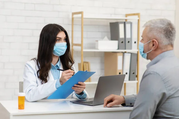 Millennial dame in beschermend masker luistert naar klachten van oude patiënt, zit aan tafel met potje pillen en laptop — Stockfoto