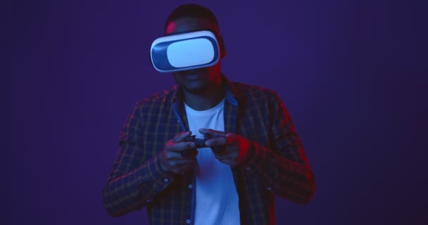 Juego VR. Joven chico negro jugando videojuegos con joystick y auriculares de realidad virtual, luces de neón de fondo — Vídeo de stock