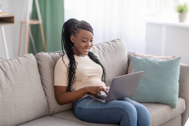 Evdeki kanepede oturan neşeli Afrikalı kadın, dizüstü bilgisayar kullanıyor.