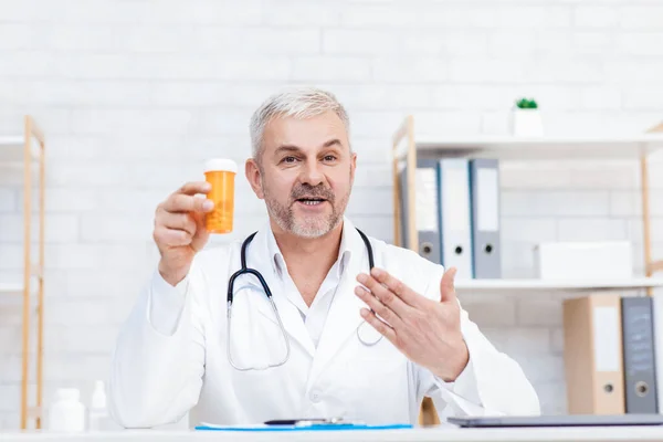 Glimlachende volwassen mannelijke arts in wit medisch uniform zit aan het bureau in het ziekenhuis, geneeskunde, gezondheidszorg — Stockfoto