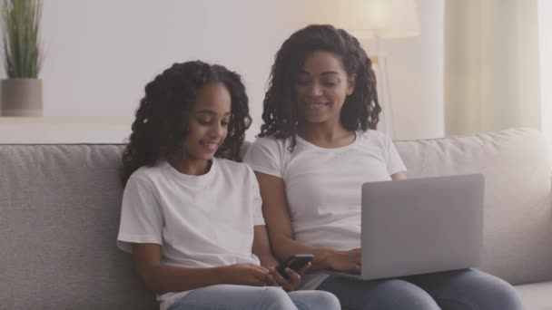 Jonge moeder en dochter zitten samen op bank en web surfen - vrouw op laptop en meisje op smartphone — Stockvideo