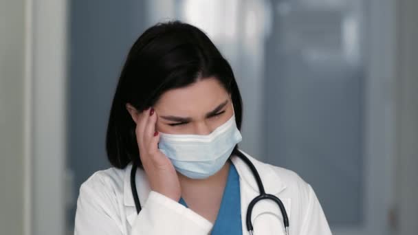 De cerca retrato médico femenino con máscara protectora que sufre de dolor de cabeza, masajeando sus sienes en el hospital — Vídeos de Stock