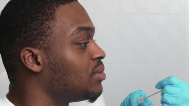 Врач в защитной маске берет образец горла ПЦР для теста Covid-19 у африканского пациента из Америки в клинике — стоковое видео