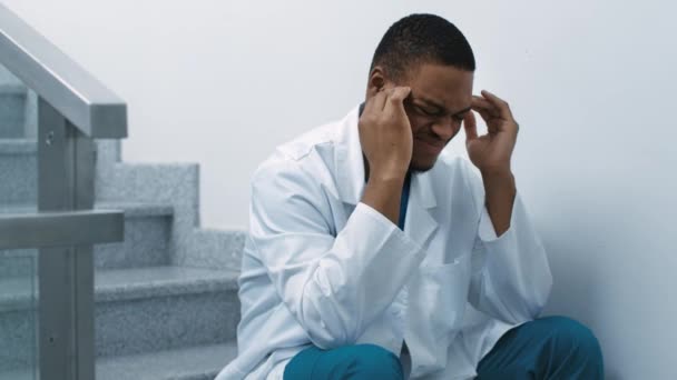 Стресс медицинских работников во время COVID-19. Уставший черный врач, сидящий на лестнице в больнице, страдающий головной болью — стоковое видео