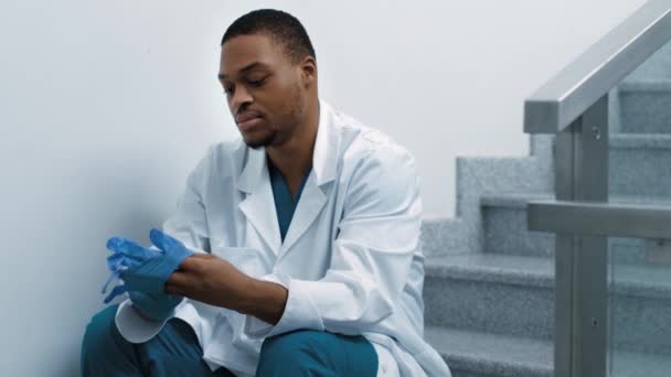 Médico afroamericano cansado descansando en las escaleras en el hospital, poniéndose guantes protectores antes del trabajo — Vídeos de Stock