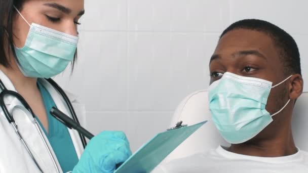 Жінка-лікар в медичній масці запитує пацієнта чорного чоловіка про симптоми, пишучи особисту історію хвороби в клініці — стокове відео