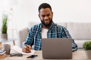 Laptop 'ta çalışan siyahi iş adamı ofise not alıyor.