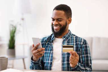 Smartphone üzerinden alışveriş yapan siyahi bir adam. Kapalı alanda oturan kredi kartı.