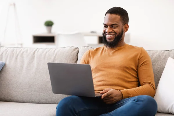 Black Man Using Laptop Computer Sitting On Couch At Home