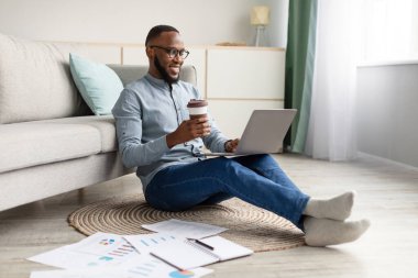 Afrikalı Adam dizüstü bilgisayar kullanıyor. Yerde oturuyor ve evde çalışıyor.