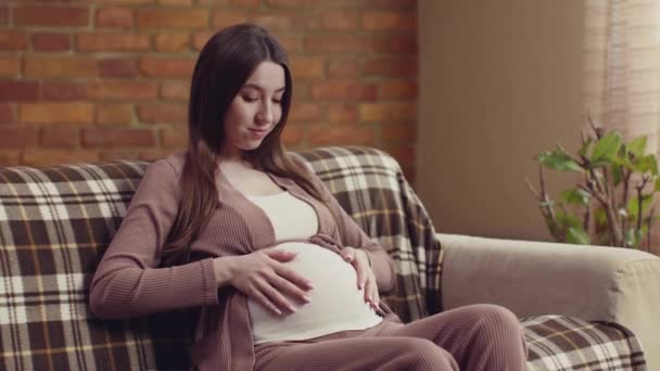 Aranyos terhes nő ül otthon a kanapén és simogatja a nagy hasát, várja, hogy találkozzon a babával, lassított felvétel — Stock videók