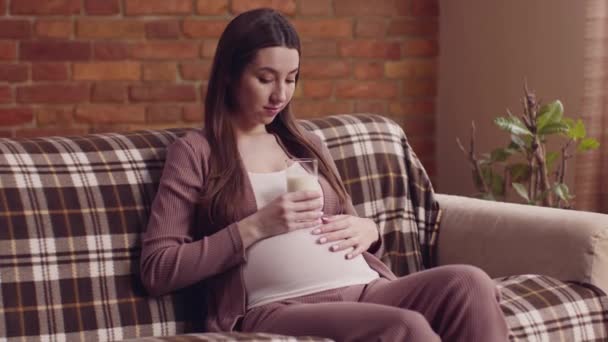 Νεαρή έγκυος γυναίκα κάθεται στον καναπέ στο σπίτι με ένα ποτήρι γάλα και χαϊδεύει τη μεγάλη κοιλιά της, αργή κίνηση — Αρχείο Βίντεο