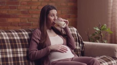 Genç hamile kadın süt içiyor, evdeki kanepede dinleniyor, kalsiyum açığını kapatıyor.
