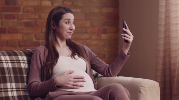 Jeune femme enceinte heureuse appel vidéo avec téléphone portable, montrant son gros ventre à la caméra, au ralenti — Video