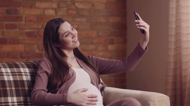 Conteúdo de mídia social. Bonito grávida senhora fazendo selfie no smartphone, se divertindo no sofá em casa — Vídeo de Stock