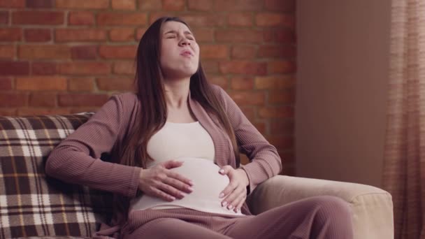 Νεαρή έγκυος γυναίκα που υποφέρει από πόνο στον τοκετό, κάθεται στον καναπέ στο σπίτι, αργή κίνηση — Αρχείο Βίντεο