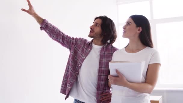 Szczęśliwi młodzi małżonkowie dyskutujący o wnętrzu nowego pustego mieszkania, kobieta trzymająca plan — Wideo stockowe
