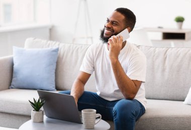 Cep telefonuyla konuşan Afrikalı adam laptopta çalışıyor, içeride oturuyor.