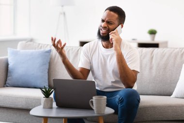 Memnuniyetsiz siyahi adam cep telefonuyla konuşuyor, bilgisayarın başında oturuyor.