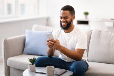 Evdeki koltukta oturan mutlu siyah adam telefonu kullanıyor.