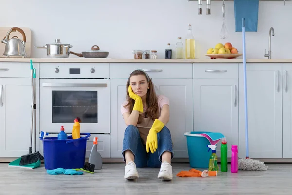 Exhausted young housewife sitting on floor at kitchen, surrounded by cleaning supplies, tired from housework