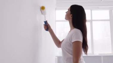 Evde tadilat yapan genç bir bayan, boyacı ve beyaz boyayla duvar boyuyor.
