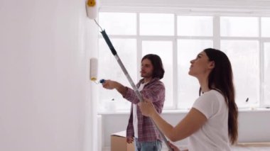 Genç evli bir çift birlikte tamirat yapıyor, boş bir dairede silindirli beyaz bir duvar boyuyorlar.