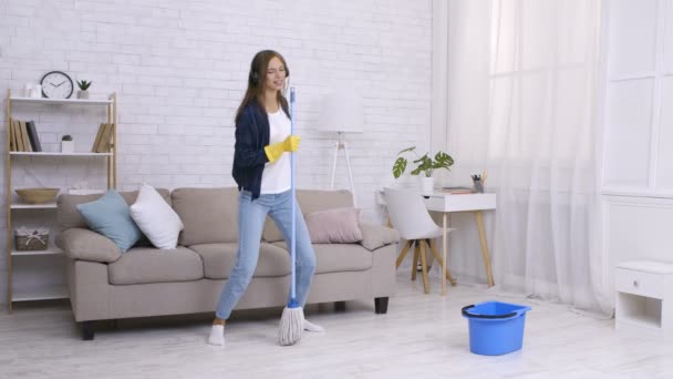 Le nettoyage printanier est amusant. Joyeux jeune femme dansant et chantant dans la serpillière comme micro, écoutant de la musique dans les écouteurs — Video