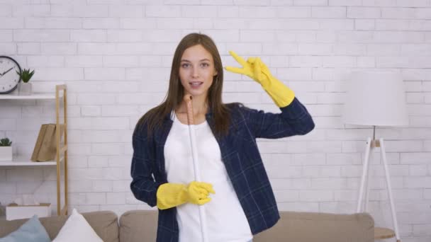 Młoda dama ciesząca się sprzątaniem domu, śpiewając do mopa jak mikrofon i tańcząc, patrząc w kamerę — Wideo stockowe
