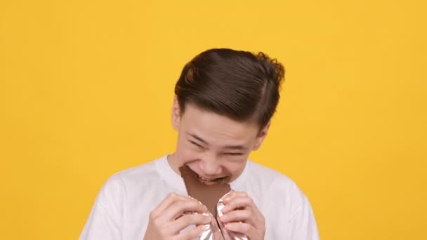 Щасливий підліток хлопчик їсть шоколад стоячи на жовтому тлі — стокове відео
