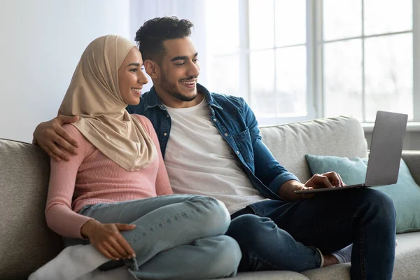 Relaxed middle-eastern couple sitting on couch, using laptop